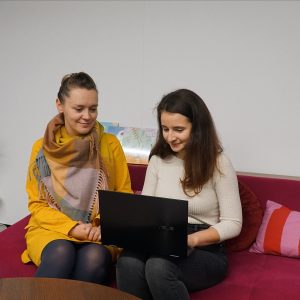 Zdjęcie przedstawia dwie kobiety siedzące na kanapie. Jedna z nich ma na kolanach laptopa.