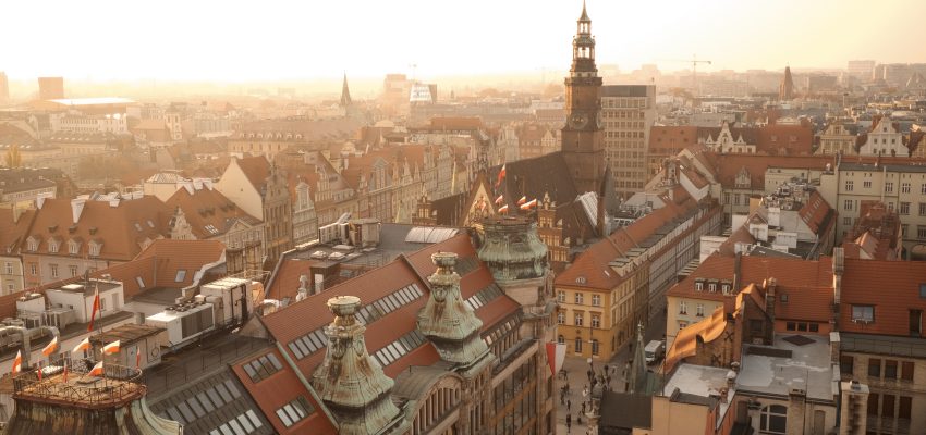 Zdjęcie przedstawia panoramę Starego Miasta we Wrocłaiu o wschodzie słońca. Na pierwszym planie widzimy dachy kamienic, w oddali wieżę Ratusza.
