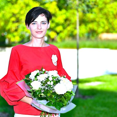 Zdjęcie przedstawia Tatianę Andrushkę w parku, trzymająca bukiet białych kwiatów i patrzącą w aparat.