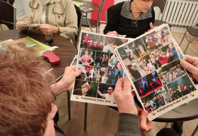 Zdjęcie przedstawia ręce dwóch osób trzymające kolaże ze zdjęć z eventów Kalejdoskop Kultur. W tle znajdują się osoby siedzące przy stole.
