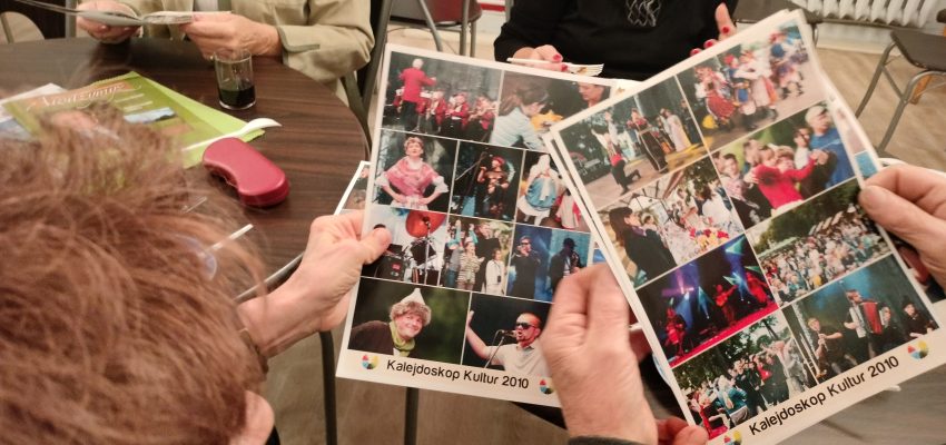 Zdjęcie przedstawia ręce dwóch osób trzymające kolaże ze zdjęć z eventów Kalejdoskop Kultur. W tle znajdują się osoby siedzące przy stole.