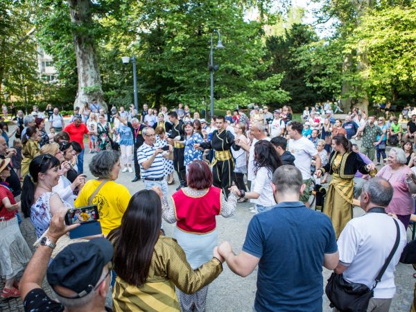 Zdjęcie przedstawia uczestników festiwalu Kalejdoskop Kultur we wspólnym tańcu w kole, gdzie osoby trzymają się za ręce.