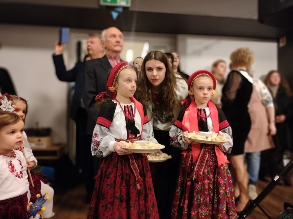 Na zdjęciu widać dwie dziewczynki ubrane na ludowo i trzymające tradycyjne dania, kobietę i kilka osób w tle.