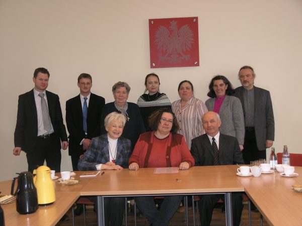 Zdjęcie przedstawia grupę osób zebraną na spotkaniu, przy stole. W pierwszym rzędzie siedzą 3 osoby, w drugim rzędzie stoi 7 osób zwróconych do aparatu. W tle na ścianie znajduje się godło Polski