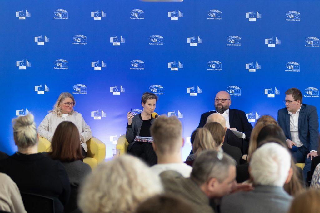 Zdjęcie przedstawia podium podczas konferecji, gdzie mówcy siedzą na fotelach. W tle grafika z logami instytucji unijnych.