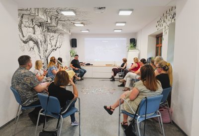 Zdjecie przedstawia sytuację szkoleniową - kilkanaście osób siedzi w kręgu, w tle 2 siedzą dwie osoby prezentujące.