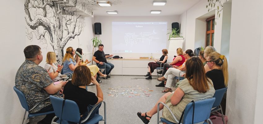 Zdjecie przedstawia sytuację szkoleniową - kilkanaście osób siedzi w kręgu, w tle 2 siedzą dwie osoby prezentujące.