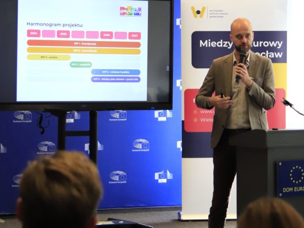 Zdjęcie przedstawia mężczyznę z mikrofonem przemawiającego na podium, na tle ekranu z prezentacją multimedialną.