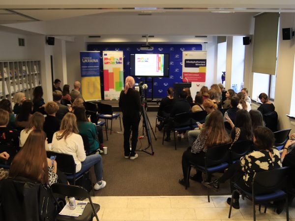 Zdjęcie przestawia wnętrze sali konferencyjnej- po dwóch stronach znajdują się siedzące osoby, na środku znajduje się kobieta obsługująca kamerę. Scena jest jeszcze pusta.