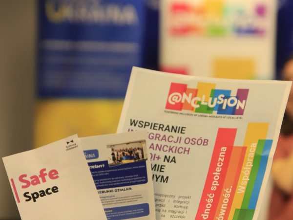 Zdjęcie przedstawia rękę, która trzyma trzy kolorowe ulotki na tle rollupów. Zdjęcie zrobione jest w taki sposób, że ostrość aparatu ustawiona jest na ulotkach, a tło jest rozmazane.