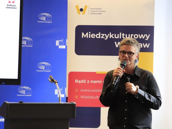 Zdjęcie przedstawia kobietę z mikrofonem przemawiająca na podium, na tle ekranu z prezentacją multimedialną.