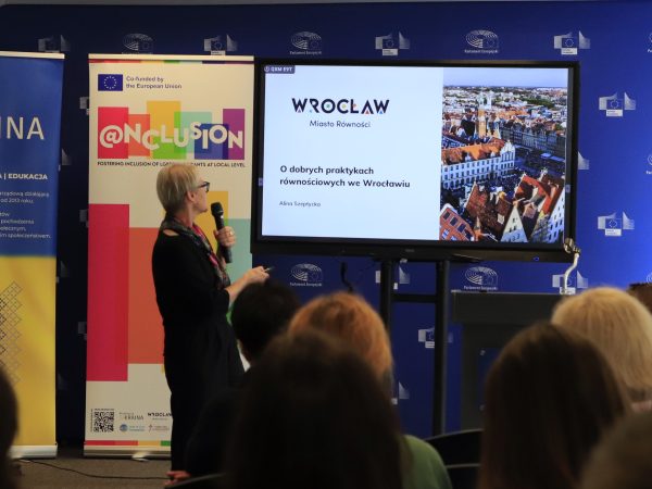 Zdjęcie przedstawia kobietę z mikrofonem przemawiająca na podium, na tle ekranu z prezentacją multimedialną.