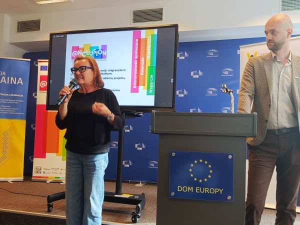 Zdjęcie przedstawia dwie osoby przemawiające na tle ekranu, z lewej strony kobieta z mikrofonem, po prawej mężczyzna.
