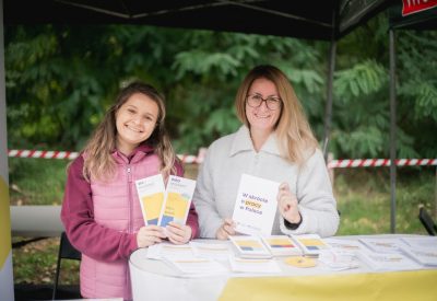 Zdjęcie przedstawia dwie uśmiechnięte kobiety na stanowisku konsultacyjnych, w rękach matrzymają i prezentują ulotki i materiały informacyjne.