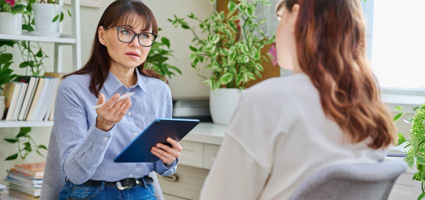 Dwie kobiety w jasnym pokoju siedzą naprzeciwko siebie - jedna z nich tyłem, a druga przodem do aparatu. Kobieta siedząca przodem ma długie ciemne włosy, okulary, ubrana jest w koszulę i dżinsy, w ręku trzyma podkładkę.