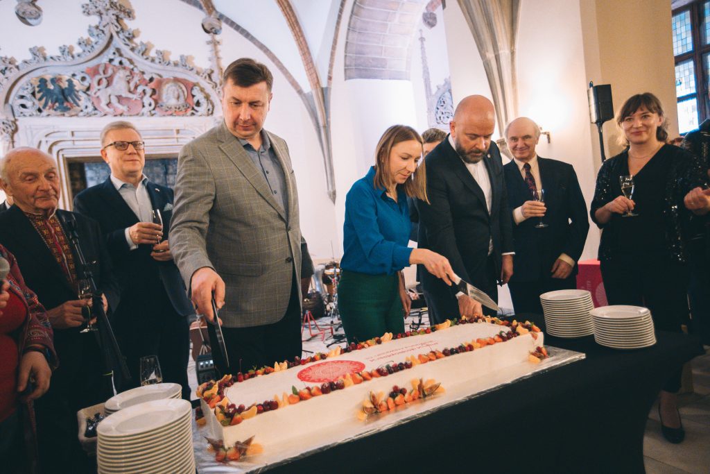 Zdjecie przedstawia mężczyznę i kobietę krojących długi, podłużny tort. Wokół nich zebrani są obserwatorzy.