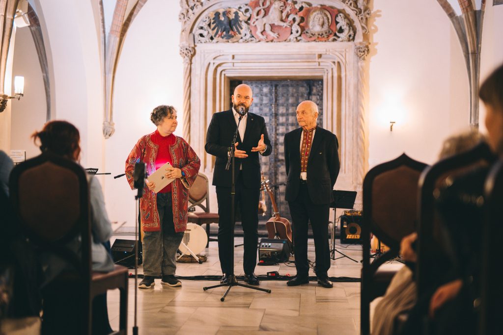 Kilkonaścioro osób w różnym wieku, ubranych elegancji, stoi na tle portalu gotyckiego we wrocławskim Ratuszu.