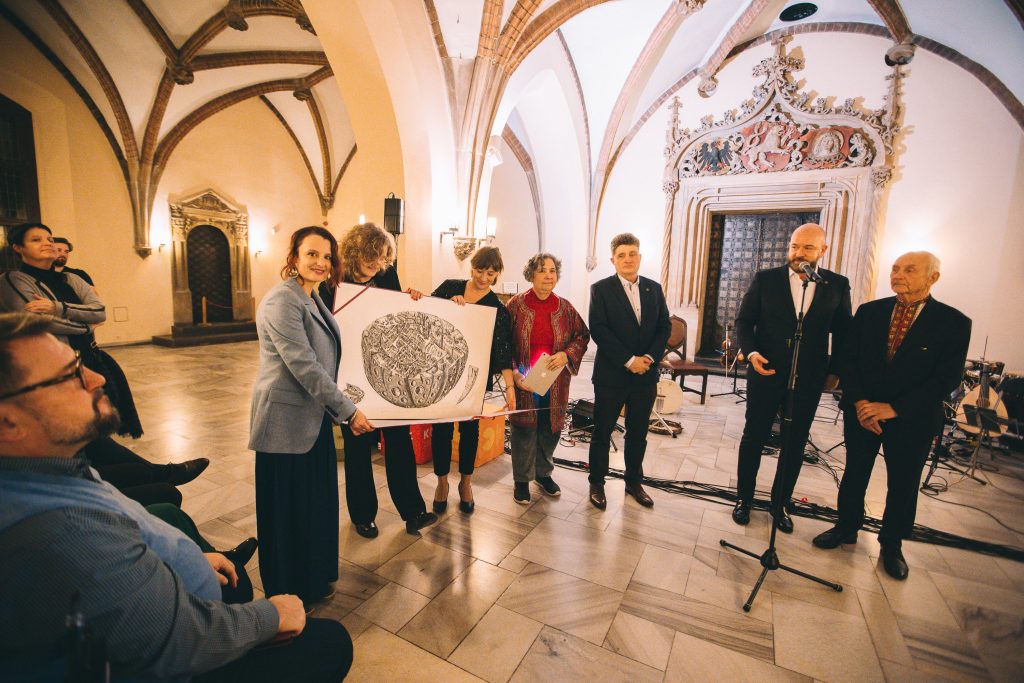 Kilkonaścioro osób w różnym wieku, ubranych elegancji, stoi na tle ozdobnych drzwi we wrocławskim Ratuszu. Kobieta w długiej marynarce, stojąca z lewej strony trzyma obraz i patrzy w stronę aparatu.