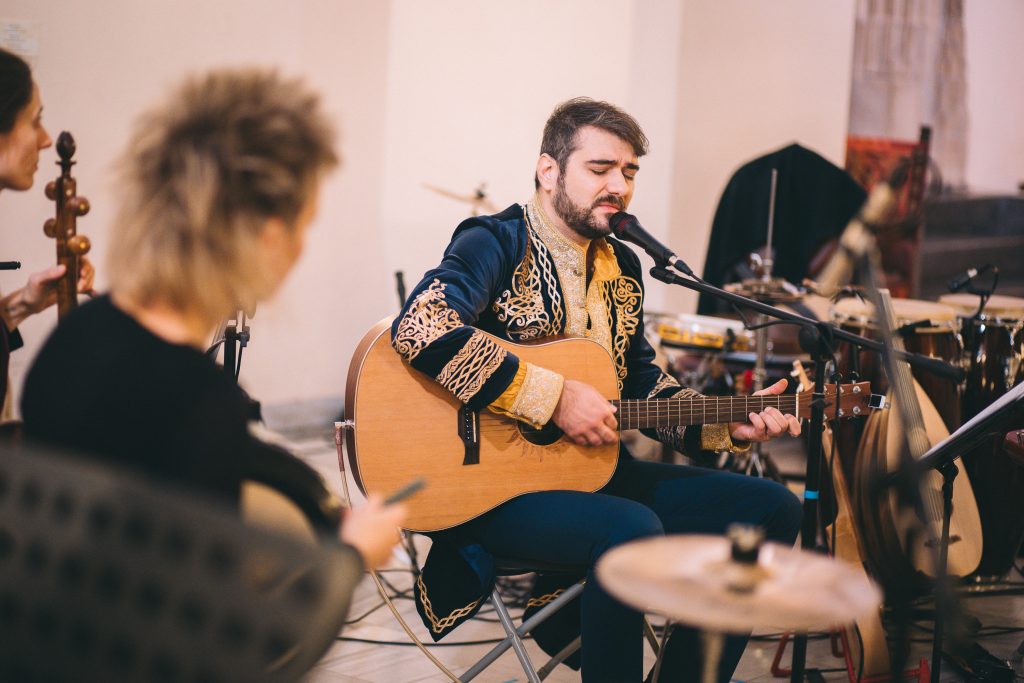 Zdjęcie przedstawia muzyków podczas koncertu, na pierwszym plane zarys kobiety, a w tle muzyk i wokalista grający na gitarze, ubrany w ozdobny, tradycyjny strój.