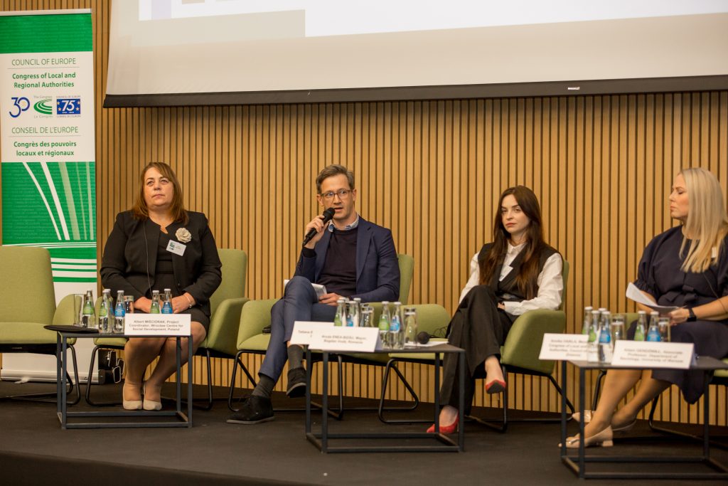 Zdjęcie przedstawia trzy kobiety i mężczyznę siedzących w fotelach na panelu dyskusyjnym. Mężczyzna w środku trzyma mikrofon i jest w trakcie wypowiedzi. Za panelistami wyświetlony jest ekran, a po lewwej stronie znajduje się roll up.