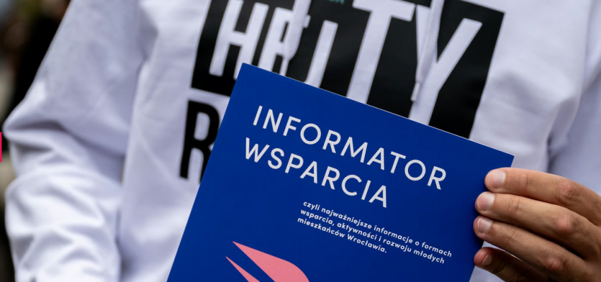 zdjęcie przedstawia tors osoby w jasne bluzie, która trzyma publikację "Informator Wsparcia"