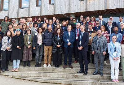 Zdjęcie przedstawia kilkadziesiąt osób stojących na betonowych schodach i uśmiechających się w stronę aparatu. W tle fragment budynku piętrowego o drewnianej elewacji i wielu oknach