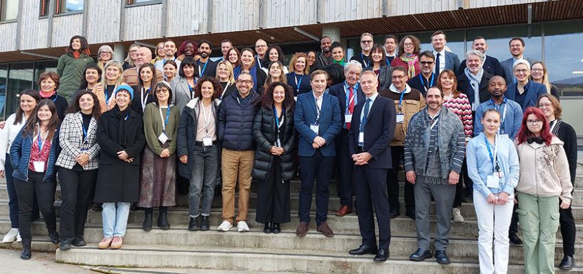 Zdjęcie przedstawia kilkadziesiąt osób stojących na betonowych schodach i uśmiechających się w stronę aparatu. W tle fragment budynku piętrowego o drewnianej elewacji i wielu oknach