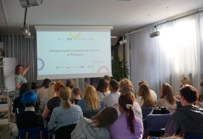 Zdjęcie przedstawia grupę osób siedzących w sali warsztatowej. Osoby sfotografowane są z tyłu. Przed nimi stoi kobieta wskazująca wyświetlaną prezentację multimedialną.