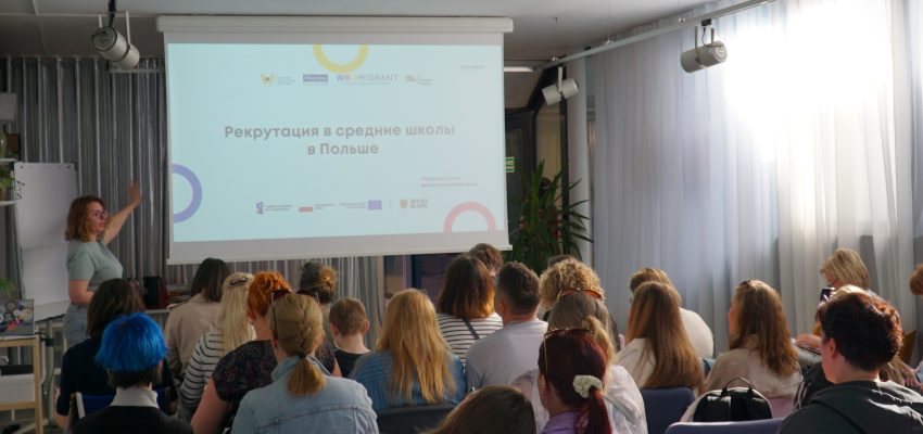 Zdjęcie przedstawia grupę osób siedzących w sali warsztatowej. Osoby sfotografowane są z tyłu. Przed nimi stoi kobieta wskazująca wyświetlaną prezentację multimedialną.