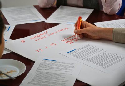 Zbliżenie na stół i rękę piszącą markerem. Wokół leżą kartki A4 z tekstem.